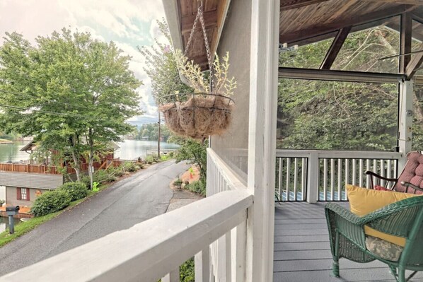 A covered porch with a view of a road and trees, a hanging plant, and a wicker chair with cushions.