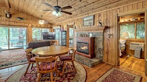A cozy cabin interior with a wooden ceiling, a fireplace, a dining table with chairs, and a television.