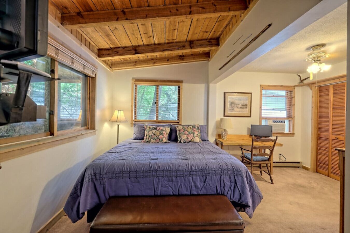 Une chambre avec un plafond en bois, un lit recouvert d’une couverture violette, un bureau avec un ordinateur et une fenêtre avec des stores.