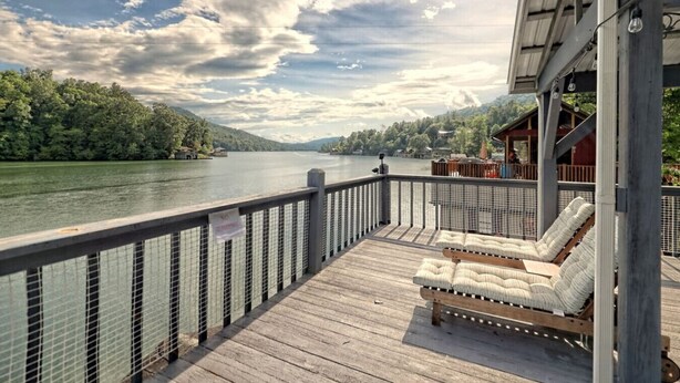 A wooden deck with a railing overlooking a lake, with a lounge chair and a covered seating area. - Lake Burton Bungalow with Top Dock & Scenic Patio (Clayton)