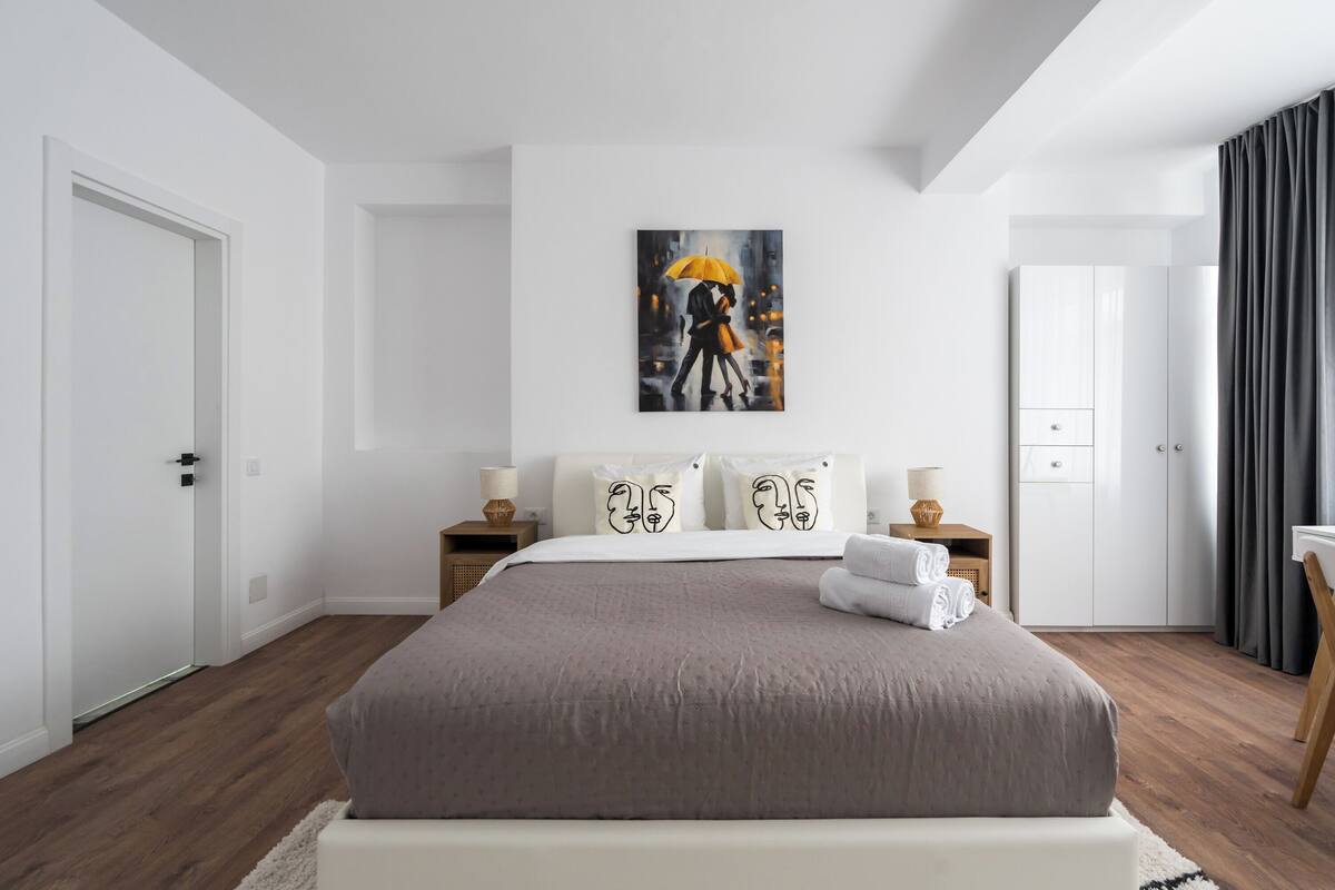 A modern bedroom with a large bed, a painting of a couple on an umbrella, white walls, and a wooden floor.