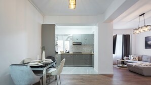 A modern kitchen with grey cabinets and a central island, a dining area with a glass table and white chairs, and a living room with a sofa and a hanging light fixture. - Pop-up Aparthotel (Bucharest)