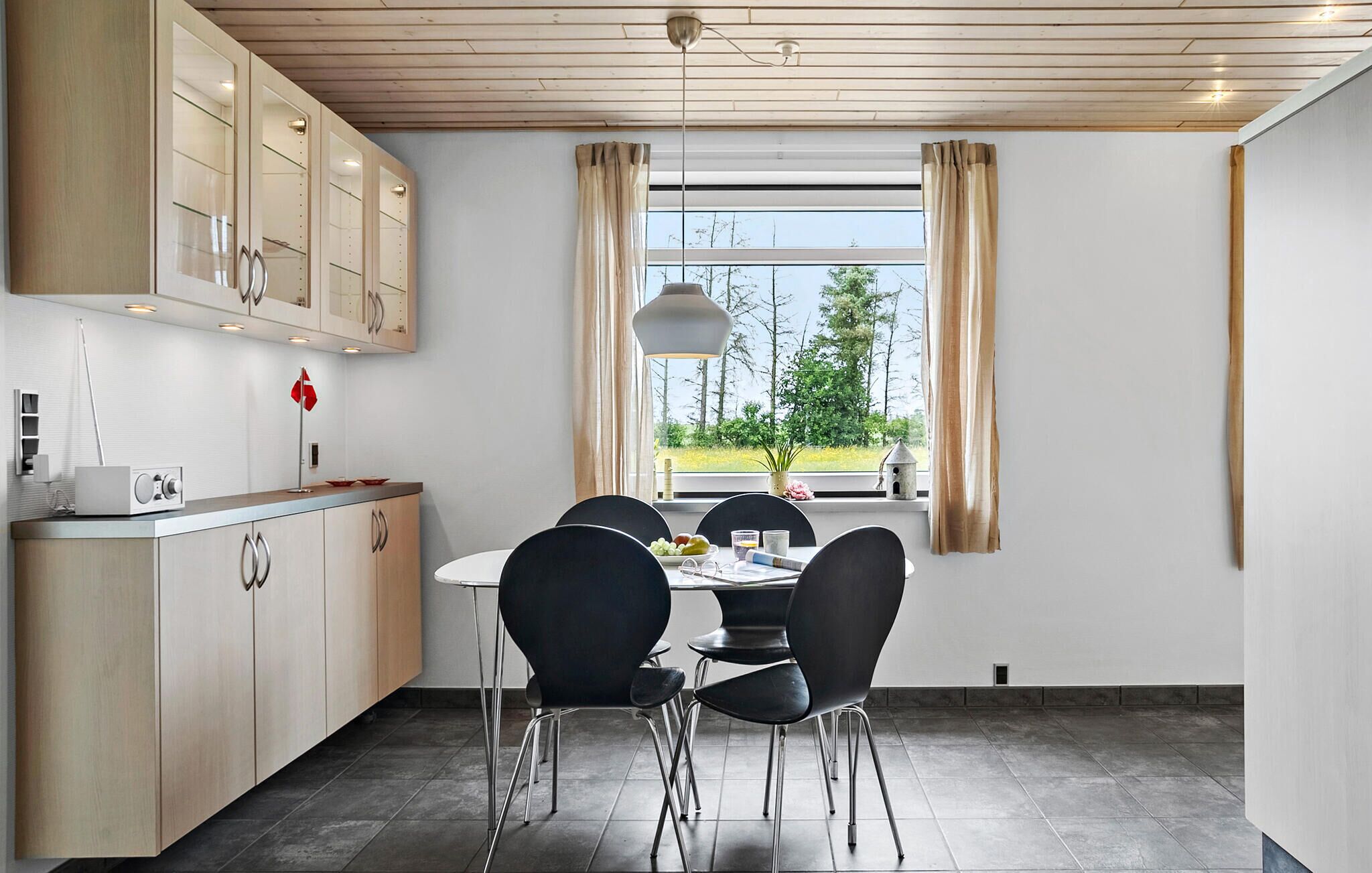 A modern kitchen with a dining area, featuring a large window with curtains, a hanging light fixture, and a view of the outdoors.
