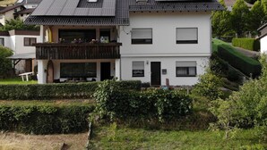 Ein zweistöckiges Haus mit Balkon, einem kleinen Garten und einem bewaldeten Hügel im Hintergrund.