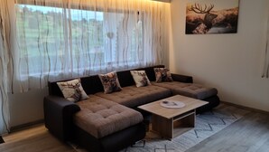 Ein modernes Wohnzimmer mit einem Ecksofa, einem Couchtisch und einem großen Fenster mit durchsichtigen Vorhängen.