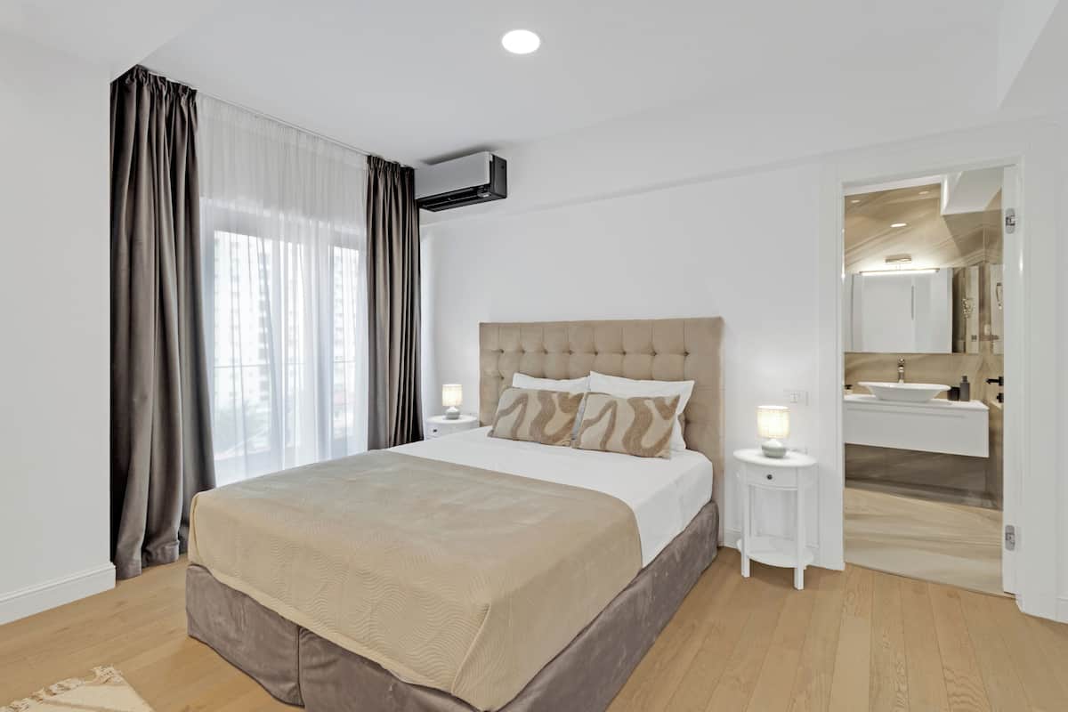 A modern bedroom with a large bed, a bedside table with a lamp, and a view of a bathroom through an open door.