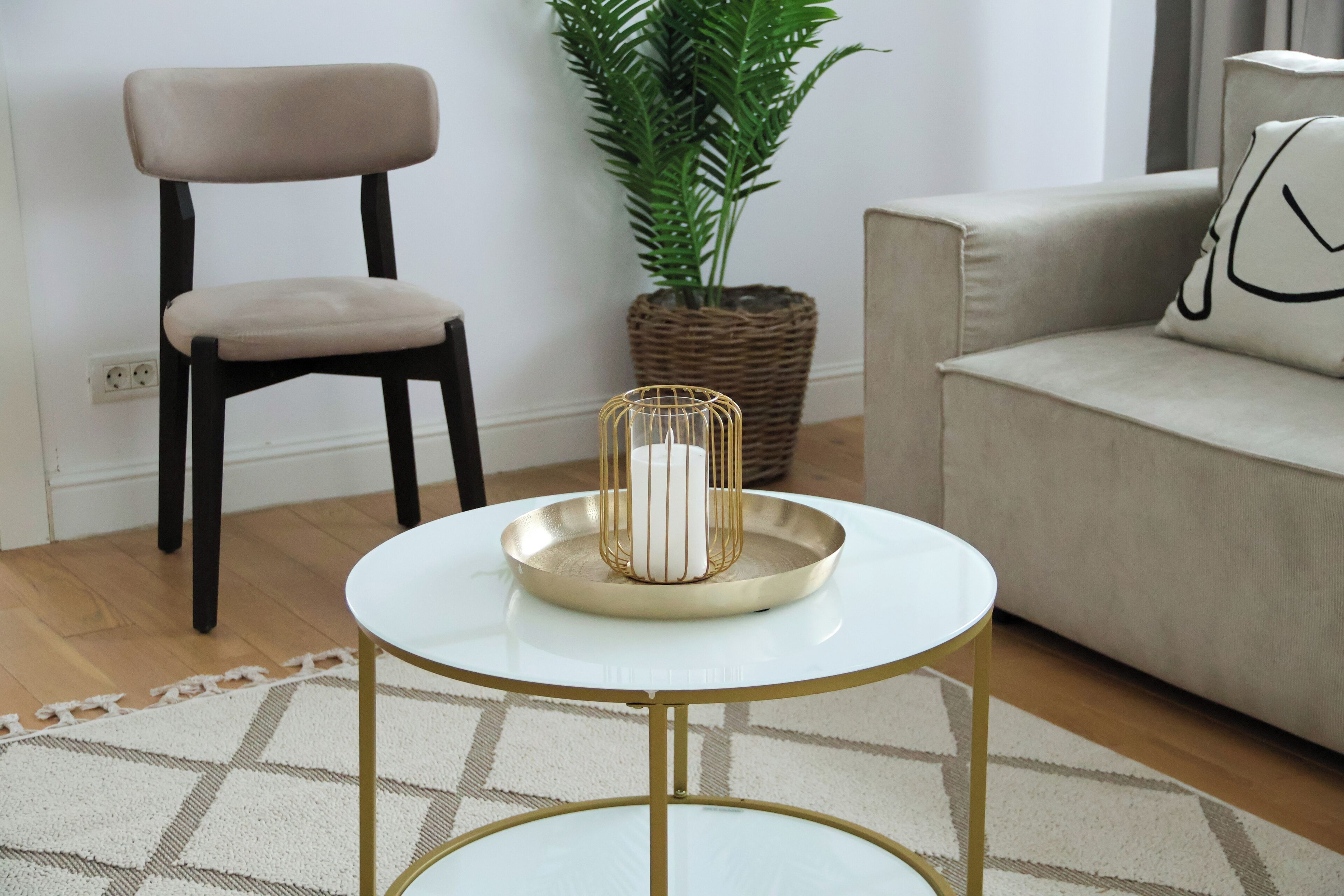 Een woonkamer met een ronde witte tafel met gouden poten, een beige stoel, een bank en een potplant.