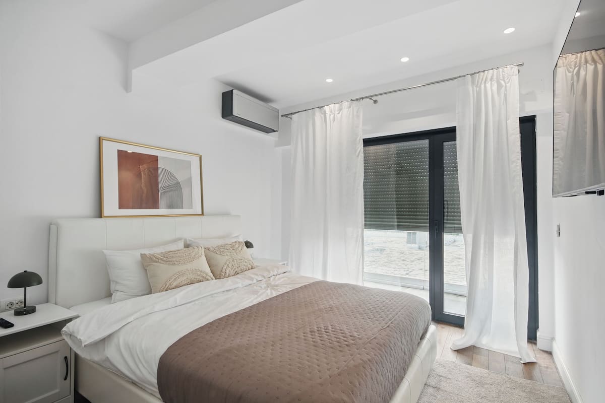A modern bedroom with a large bed, a bedside table with a lamp, a framed artwork on the wall, and a view of the beach through the window.