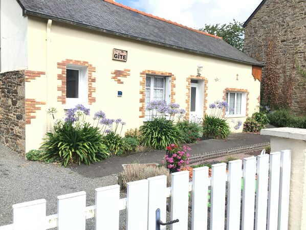 Un bâtiment sur lequel est apposée une enseigne indiquant « GÎTE », ainsi qu’un petit jardin agrémenté de fleurs et de plantes.