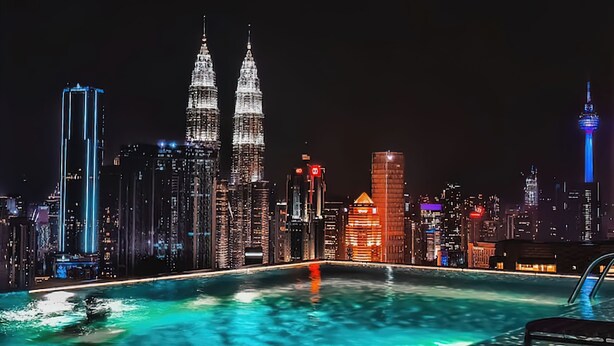 Ein beleuchtetes Stadtbild bei Nacht mit hoch aufragenden Wolkenkratzern, darunter die Petronas Towers, und einem Pool, der das Stadtlicht spiegelt.