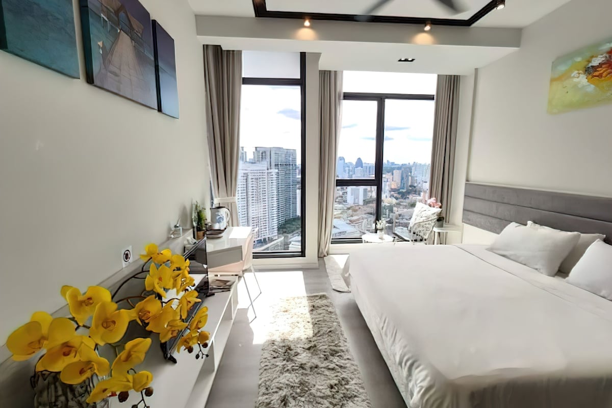 Quarto de hotel moderno com uma cama grande, uma escrivaninha e vista para a paisagem urbana através de amplas janelas.