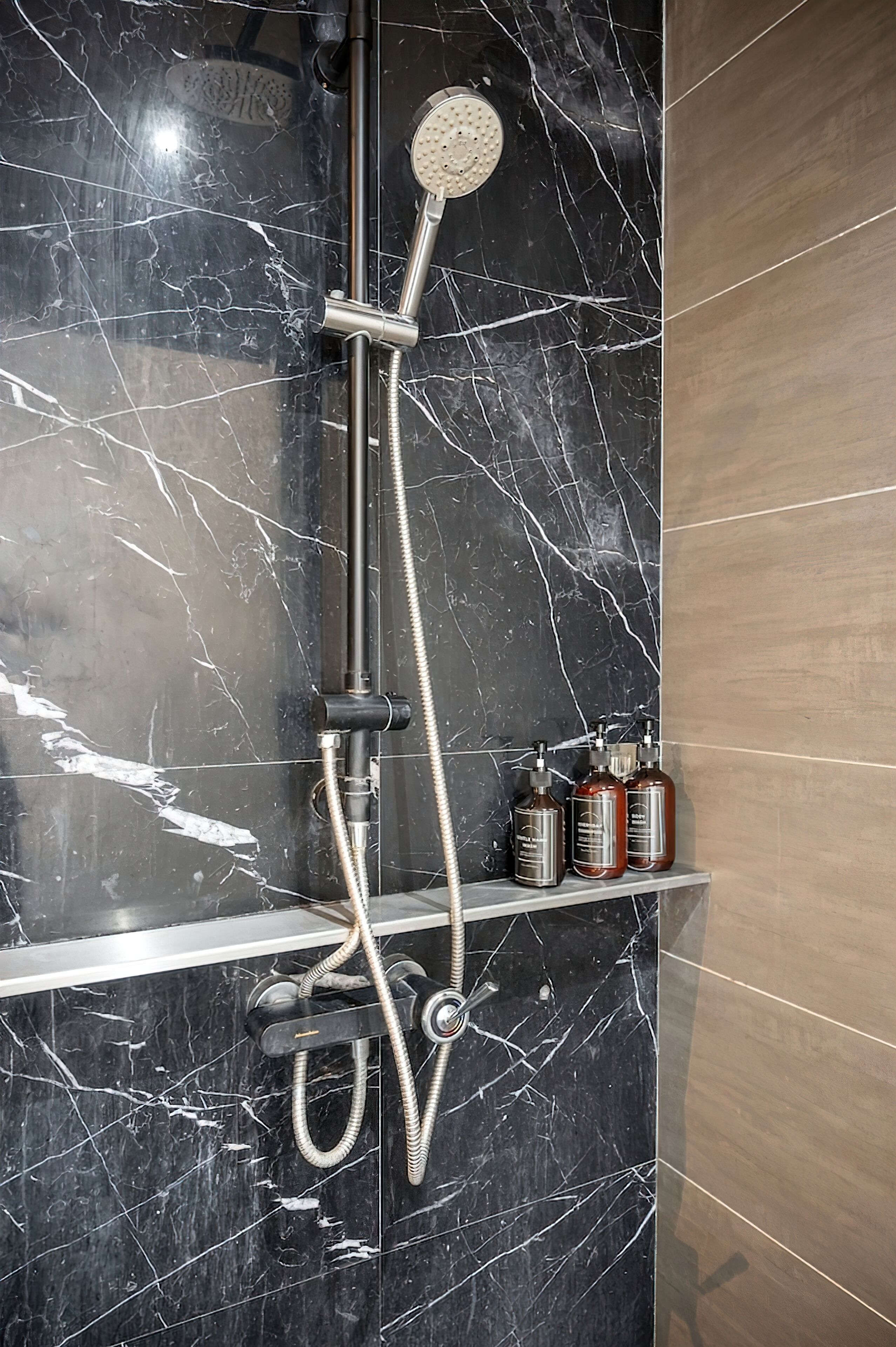 A bathroom with a black marble shower enclosure, a handheld showerhead, a wall-mounted faucet, and a shelf with toiletries.