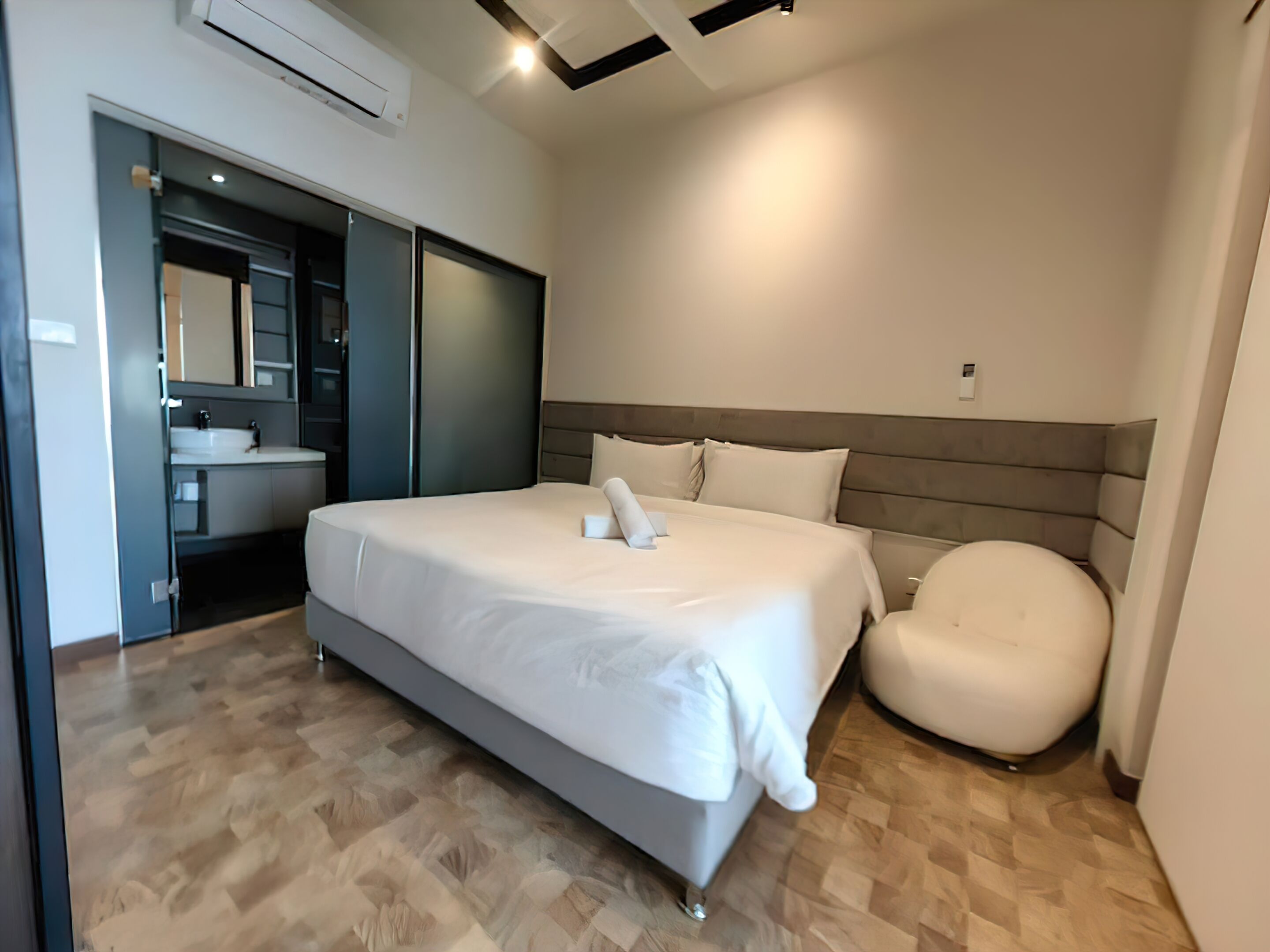 Un dormitorio moderno con una cama grande, una butaca junto a la cama, un armario empotrado y un baño con lavamanos y espejo.