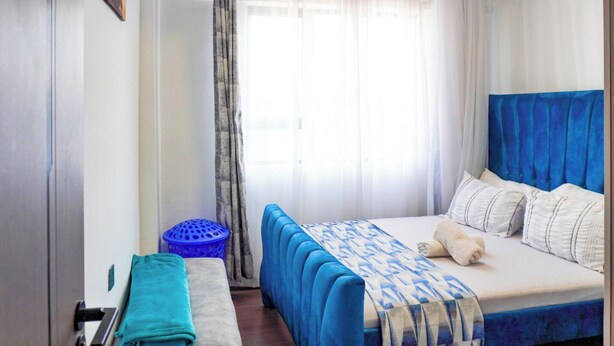 Une chambre à coucher avec un lit, une tête de lit bleue, une fenêtre avec des rideaux légers et un tableau au mur.