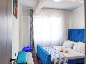 Une chambre à coucher avec un lit, une tête de lit bleue, une fenêtre avec des rideaux légers et un tableau au mur.