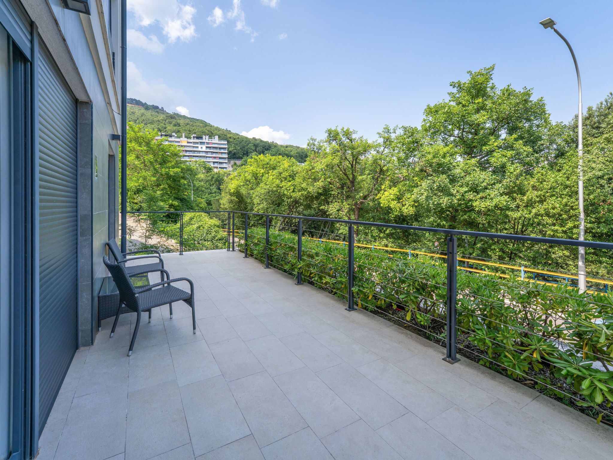A balcony with a chair, a metal railing, and a view of greenery and buildings in the background.