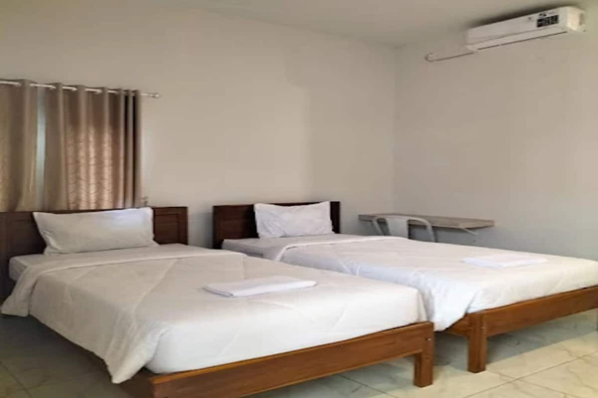 A hotel room with two single beds, a small table, a chair, and an air conditioning unit.