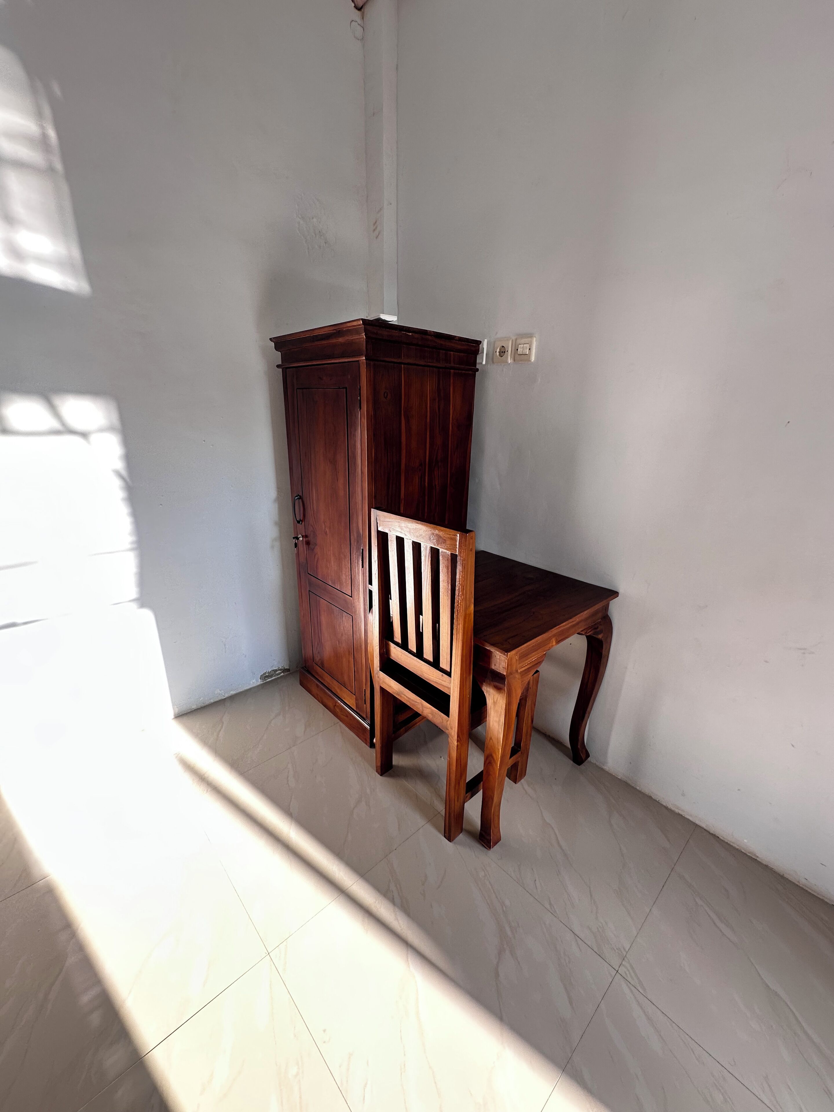 Ein Eckzimmer mit Holzschrank und Stuhl, weißen Wänden und Fliesenboden.