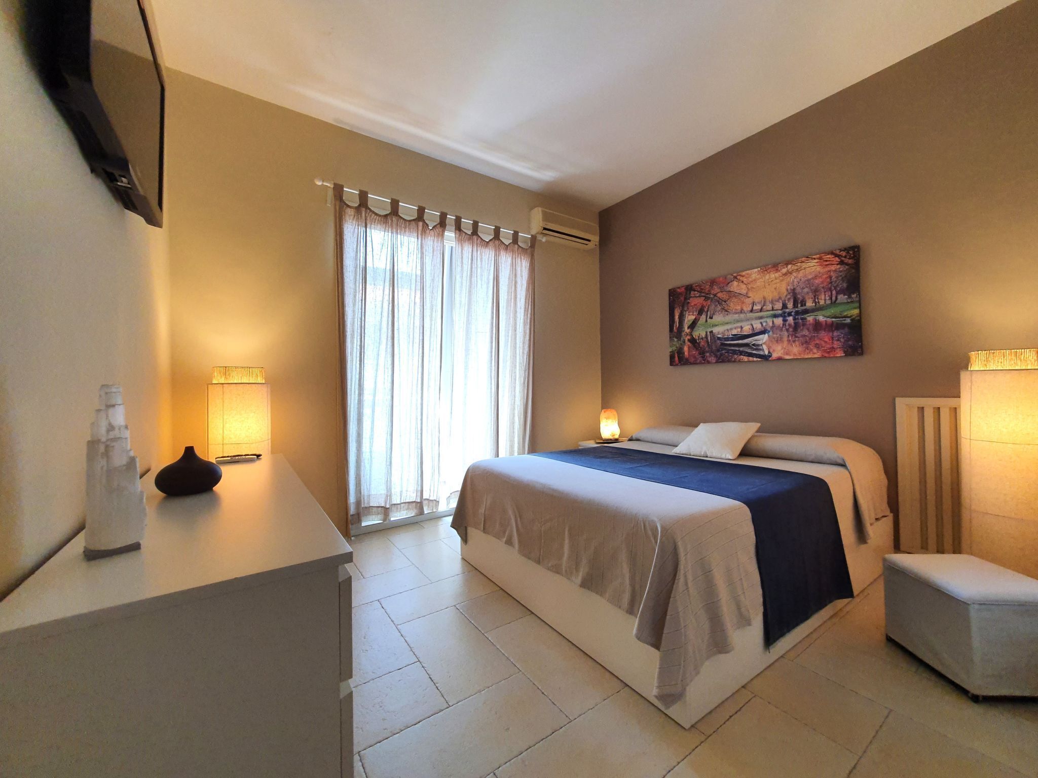 Ein Hotelzimmer mit einem Bett, Nachttischlampen, einem Gemälde und einem Schreibtisch mit einer Vase.