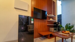 A modern hotel room with a wooden partition, a flat-screen TV, a refrigerator, a small table with fruits, and a plant.