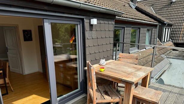 A wooden outdoor dining area with a table and benches, a sliding glass door leading to an interior space, and a tiled roof. - Saarner Loft Apartment with Private Terrace, Garden, and Wi-Fi (Mülheim an der Ruhr)