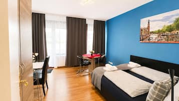 A hotel room with a bed, a desk with a chair, a window with curtains, and a painting on the wall.