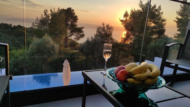 A table with a glass of wine and a bowl of fruit, overlooking a scenic view of trees and a sunset.