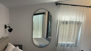 A neatly made bed with white linens, a bedside lamp, a large mirror, and a window with sheer curtains.