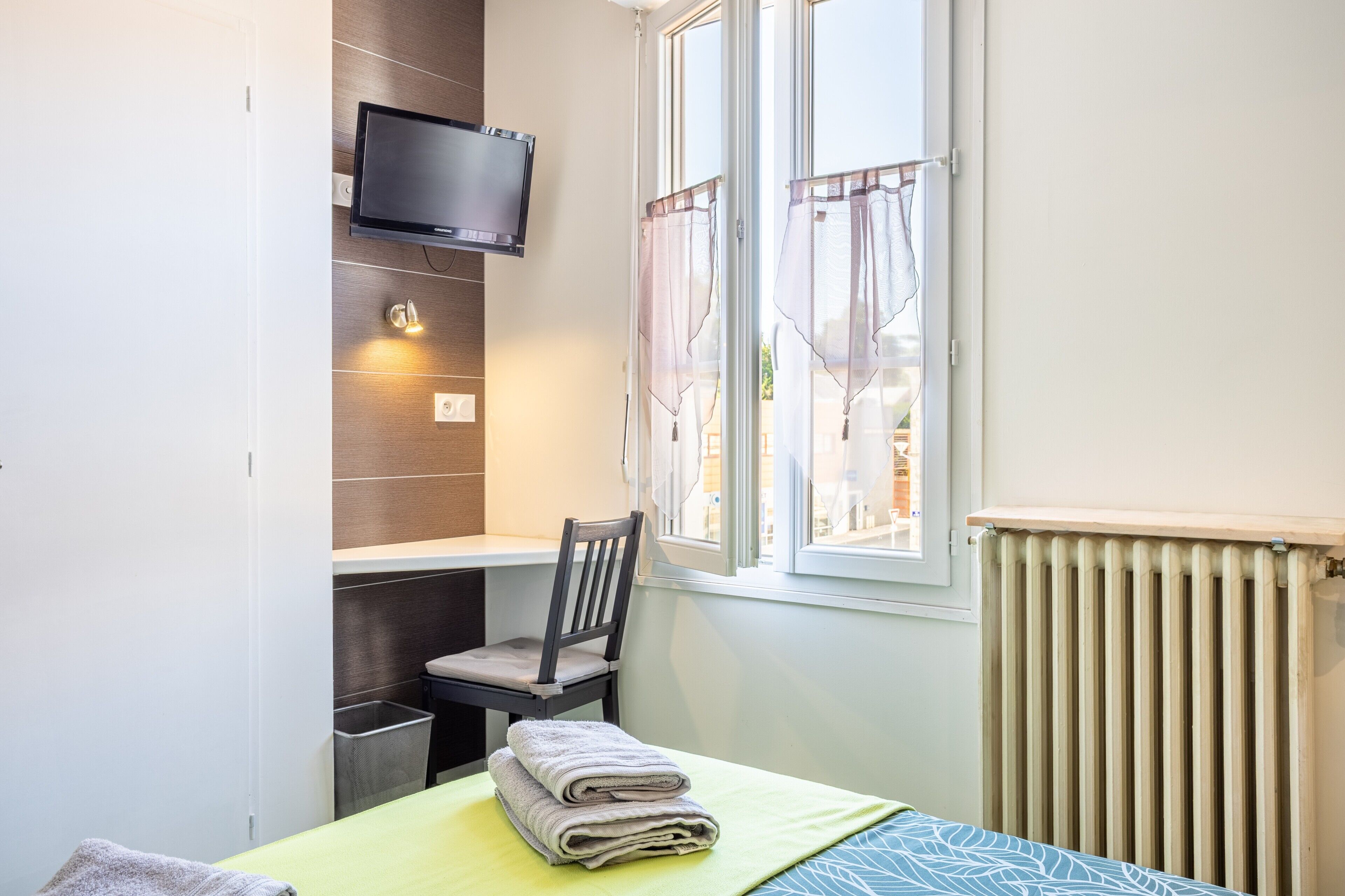 A room with a bed, a chair, a TV mounted on the wall, a window with curtains, and a radiator.