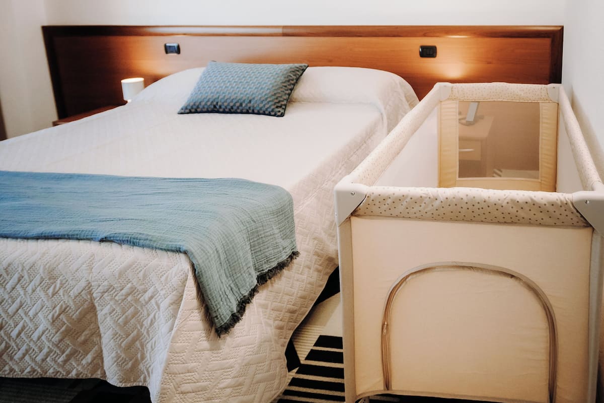 A hotel room with a double bed, a side table with a lamp, a crib, and a striped rug.