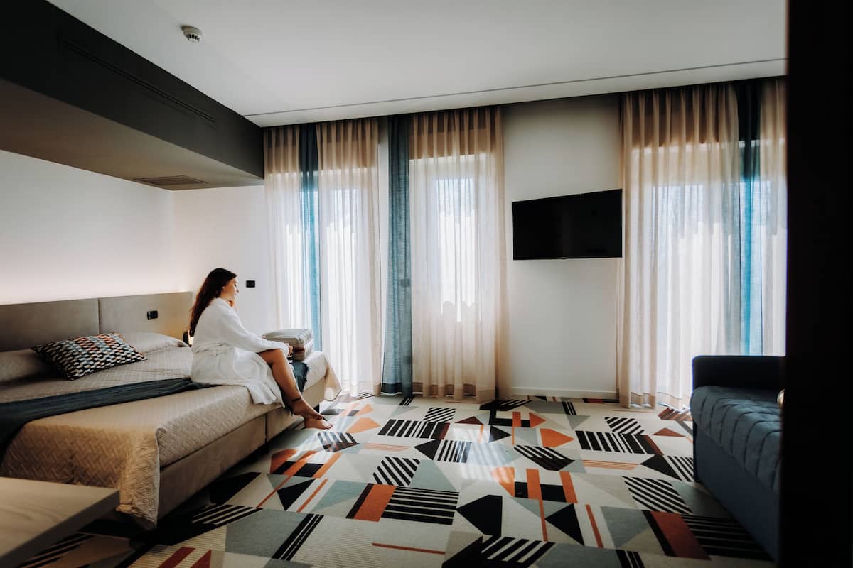 A modern hotel room with a large bed, a sofa, a TV mounted on the wall, and a patterned carpet.