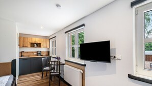 Ein kompakter Raum mit Bett, Esstisch und Stühlen, einer Küchenzeile mit Holzschränken, einem an der Wand montierten Fernseher und einem Fenster mit Blick ins Grüne.