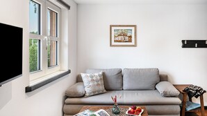 Ein Wohnzimmer mit einer Couch, einem Couchtisch mit Früchten und einer Vase, einem gerahmten Bild an der Wand und einem an der Wand befestigten Fernseher.