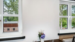Ein Zimmer mit einem Holztisch, zwei Fenstern mit Blick auf Bäume und ein Gebäude sowie einer Vase mit Blumen auf der Fensterbank.