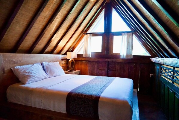 A bedroom with a wooden ceiling, a bed with white linens, a nightstand with a lamp, and a window with curtains.