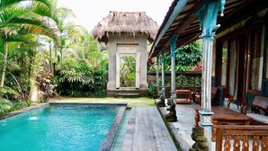 A pool with a thatched-roof structure in the background, surrounded by lush greenery and wooden furniture.