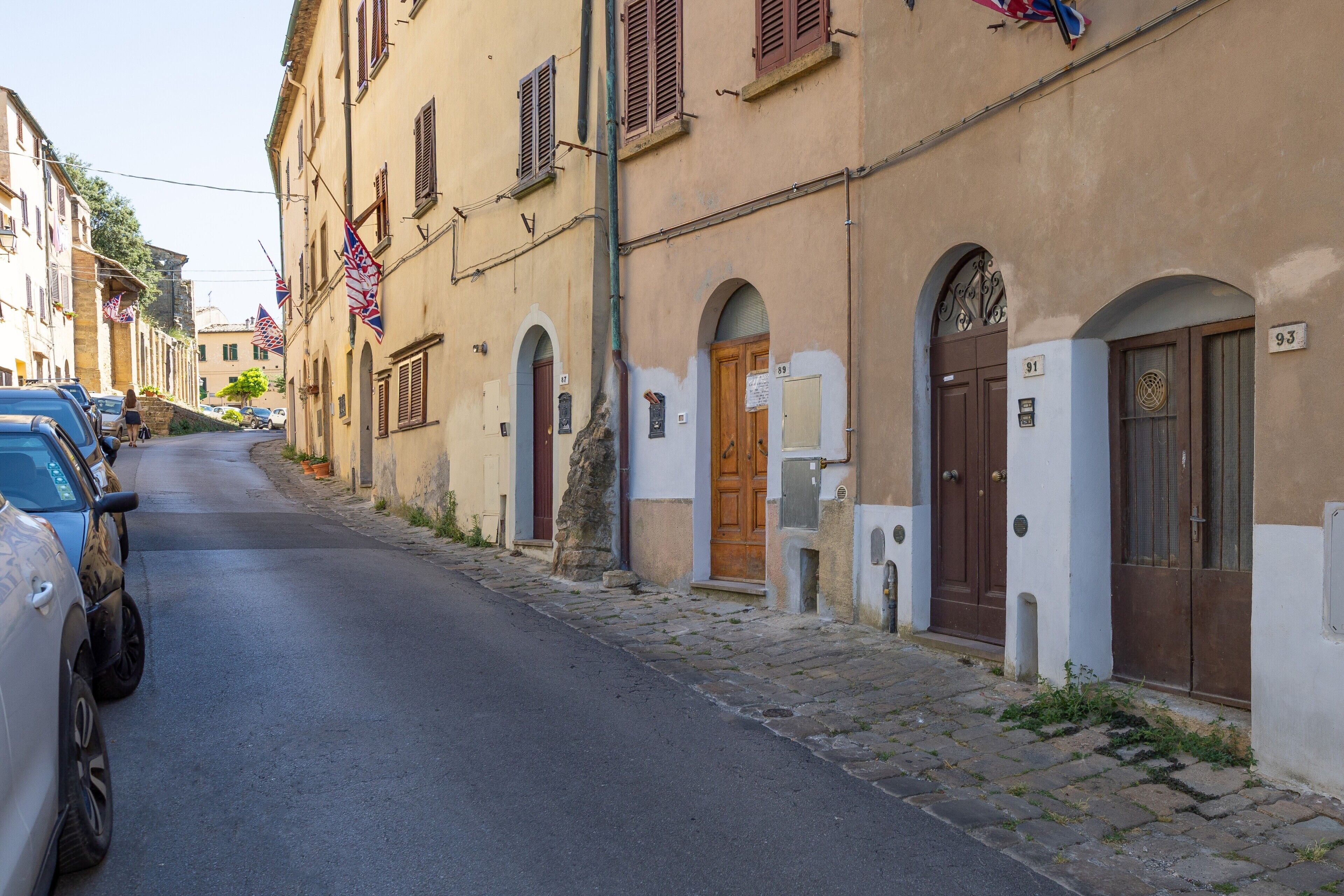 Une rue étroite bordée de bâtiments historiques, dont certains présentent des portes et des fenêtres en arc de cercle, et où quelques voitures sont garées le long du trottoir.