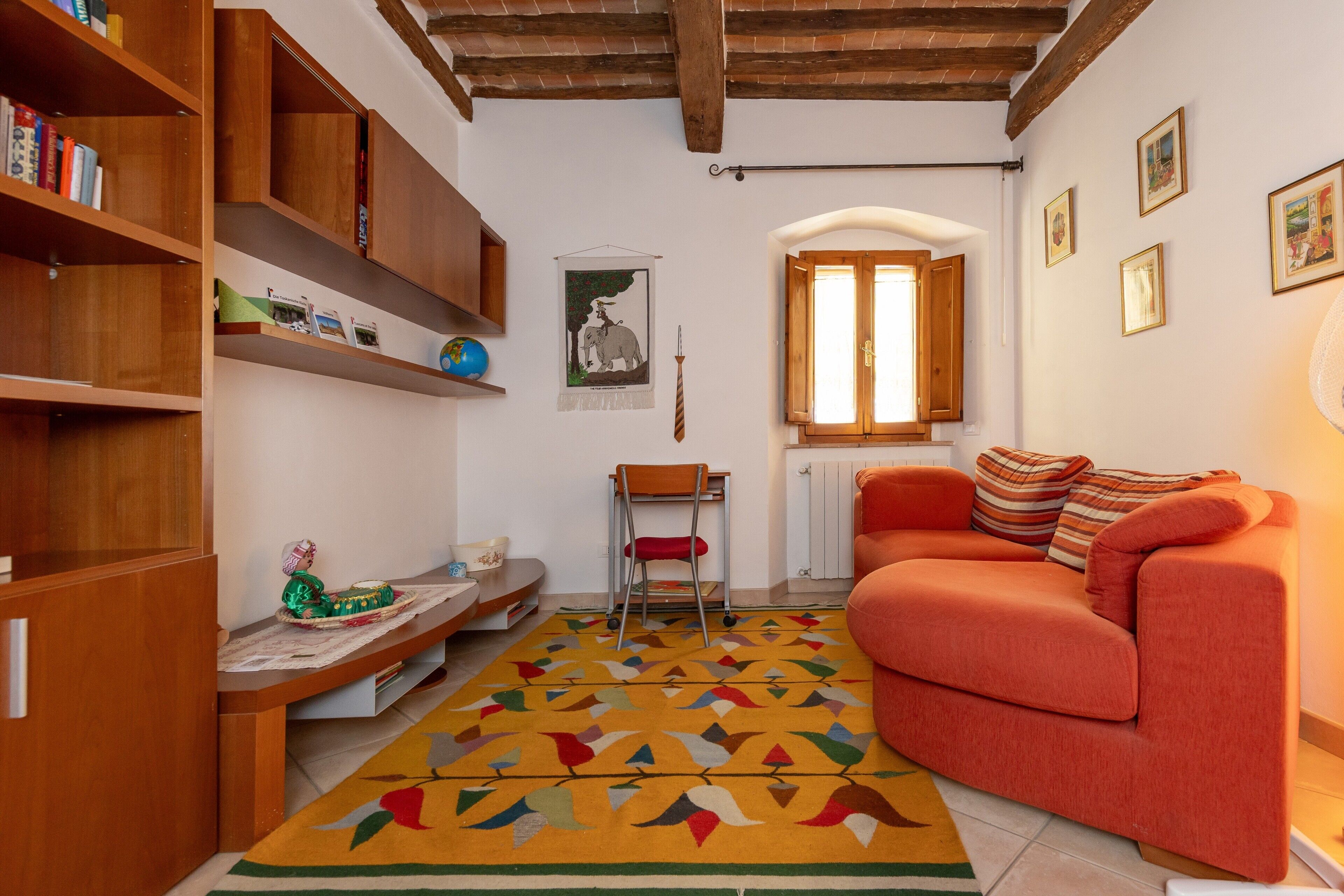Una sala de estar acogedora con un sofá, una estantería de madera, un tapete estampado y una ventana con contraventanas de madera.