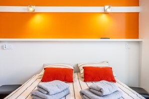 Un lit double avec une literie à rayures et des oreillers rouges, une tête de lit blanche et un mur d’accent d’un orange vif.
