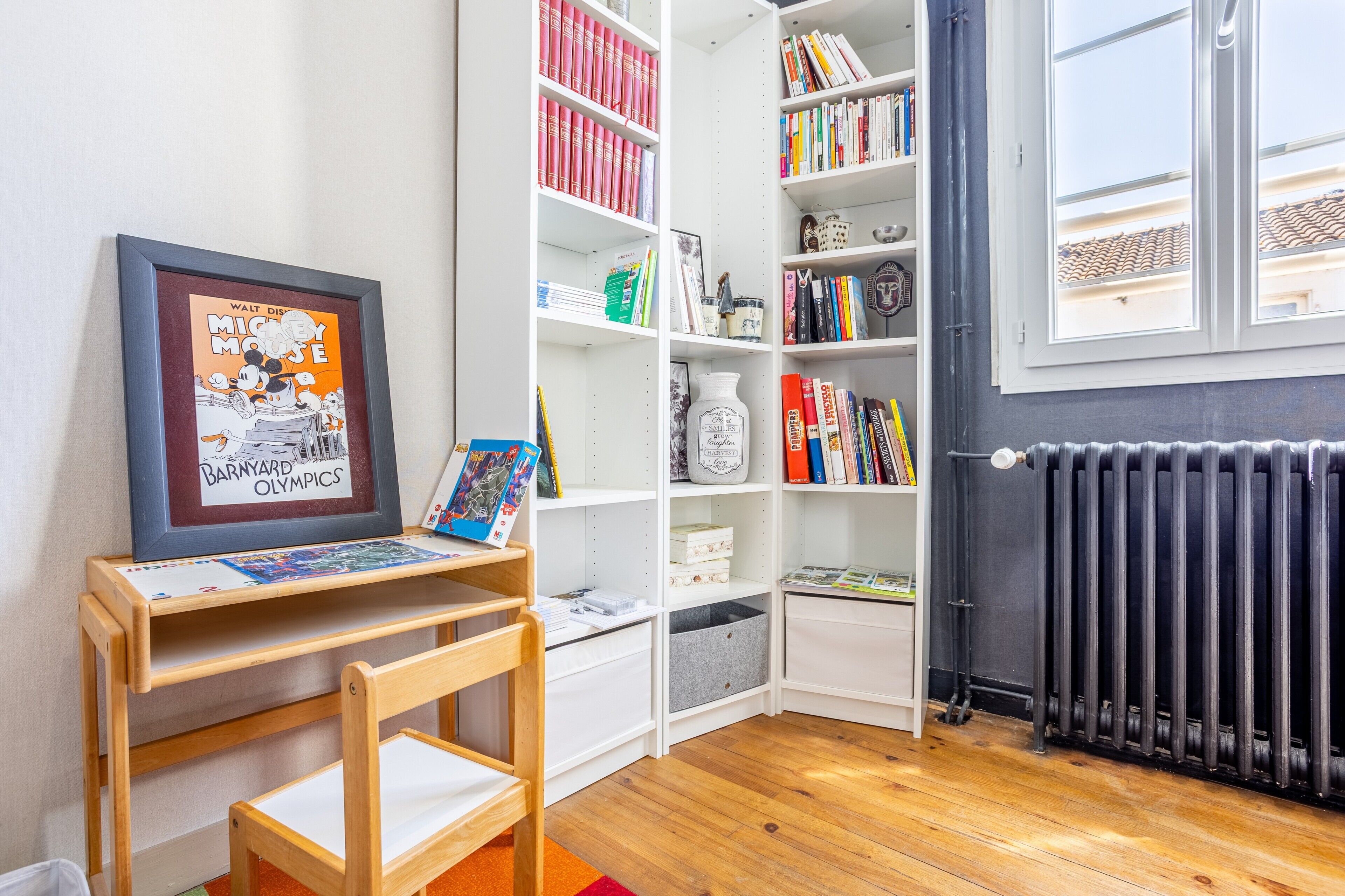 A room with a bookshelf, a framed picture, a radiator, and a window.