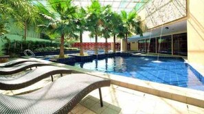 An indoor swimming pool with a tiled floor, surrounded by palm trees and a covered area with a glass ceiling. - Panoramic Skyline Views Cozy Vibes Near It All (Mandaluyong City)