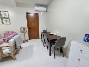 A compact living space with a dining table, chairs, a fan, a small cabinet, and a door. - Panoramic Skyline Views Cozy Vibes Near It All (Mandaluyong City)