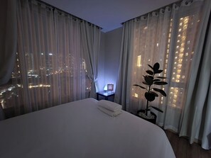 A hotel room with a large bed, a nightstand with a lamp, and a view of a cityscape through sheer curtains. - Panoramic Skyline Views Cozy Vibes Near It All (Mandaluyong City)