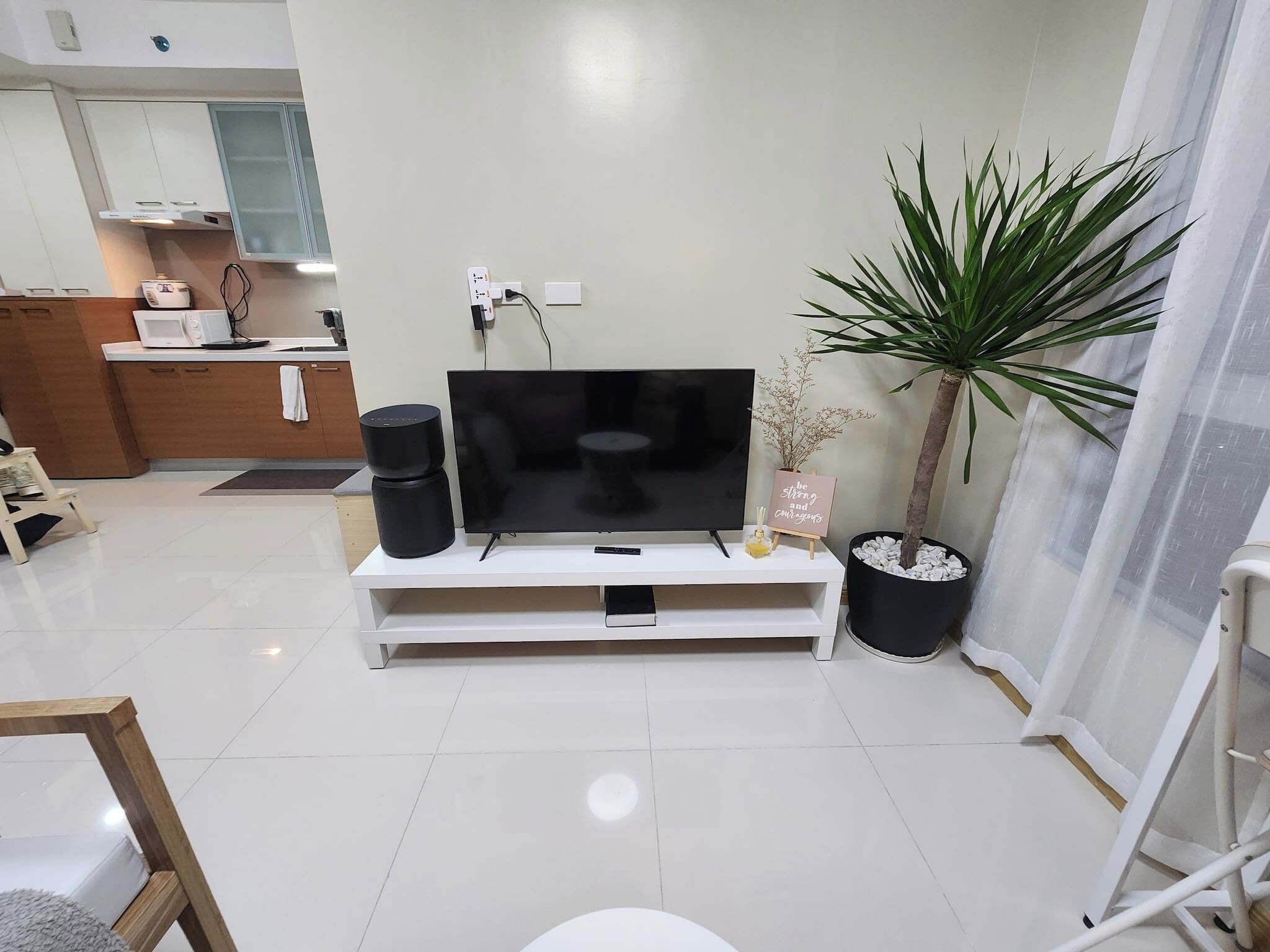 A modern living room with a flat-screen TV on a white stand, a potted plant, and a small table with a sign.