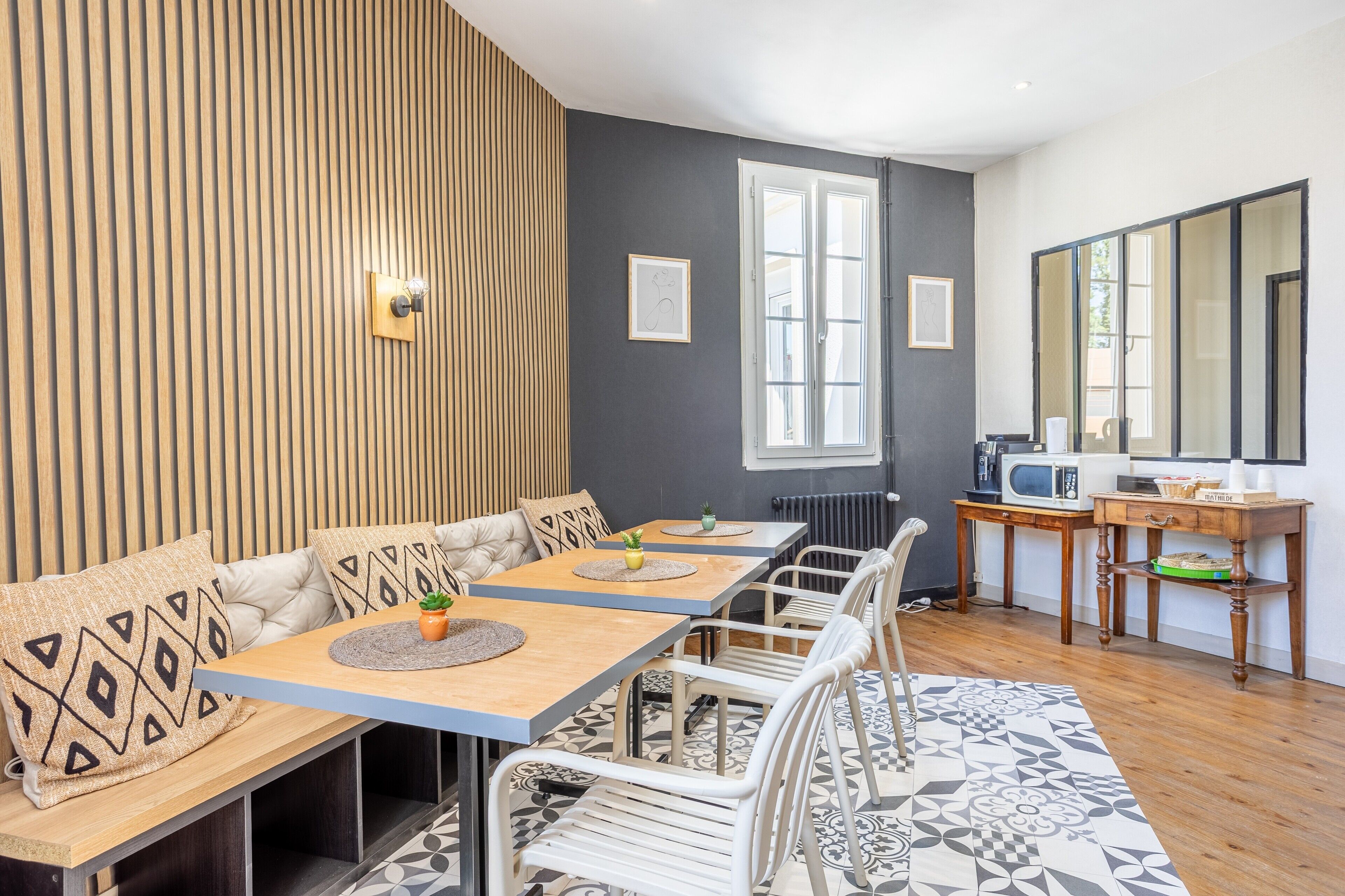 A dining area with a long table, chairs, and a bench with cushions. There is a wooden wall, a window, and a microwave on a table.