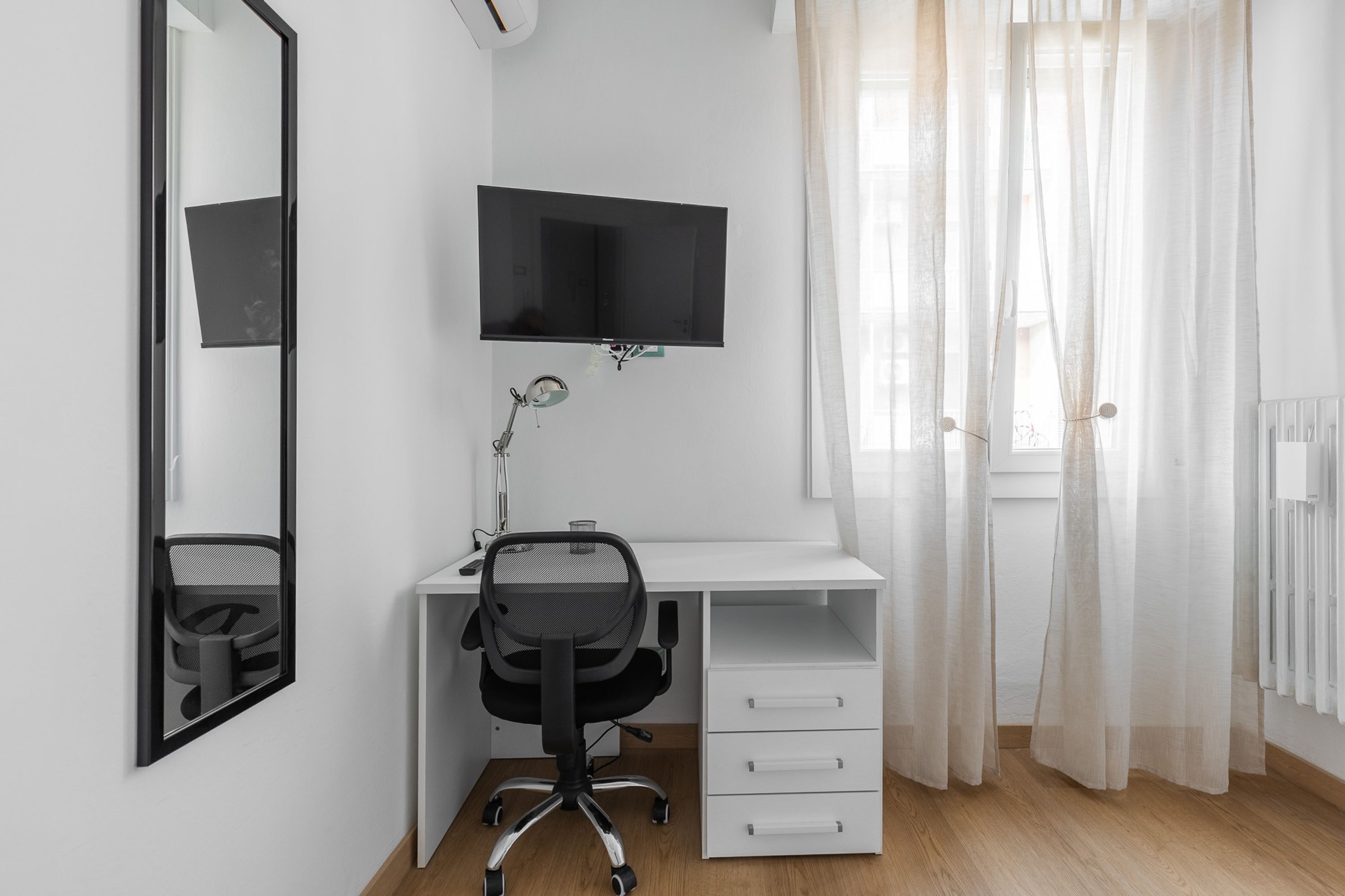 A modern, minimalist bedroom with a white desk, a black office chair, a mounted TV, and a window with sheer curtains.