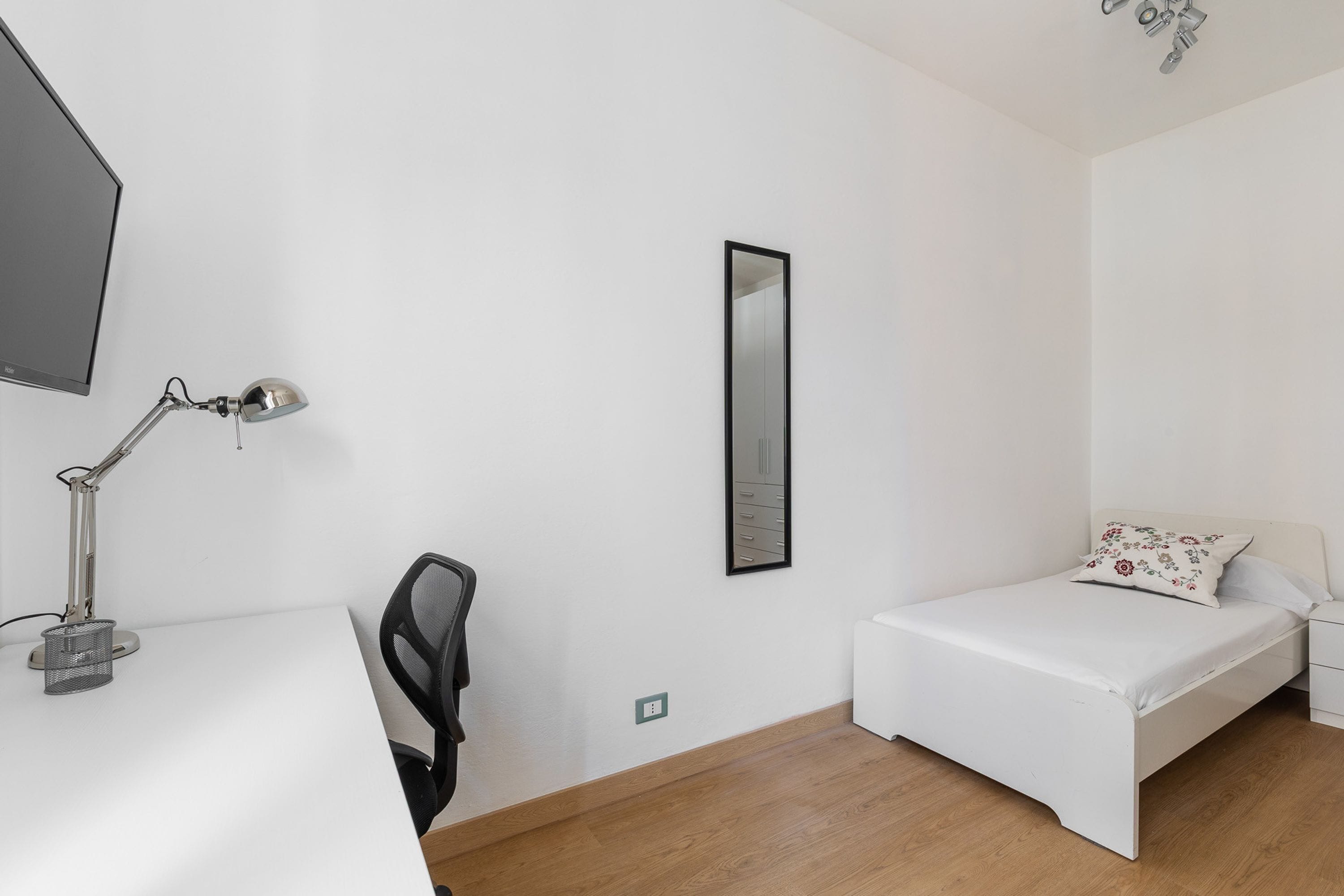 Una habitación pequeña y limpia con un escritorio blanco, una silla, una cama con colchón blanco y un cojín floral, y un espejo en la pared.