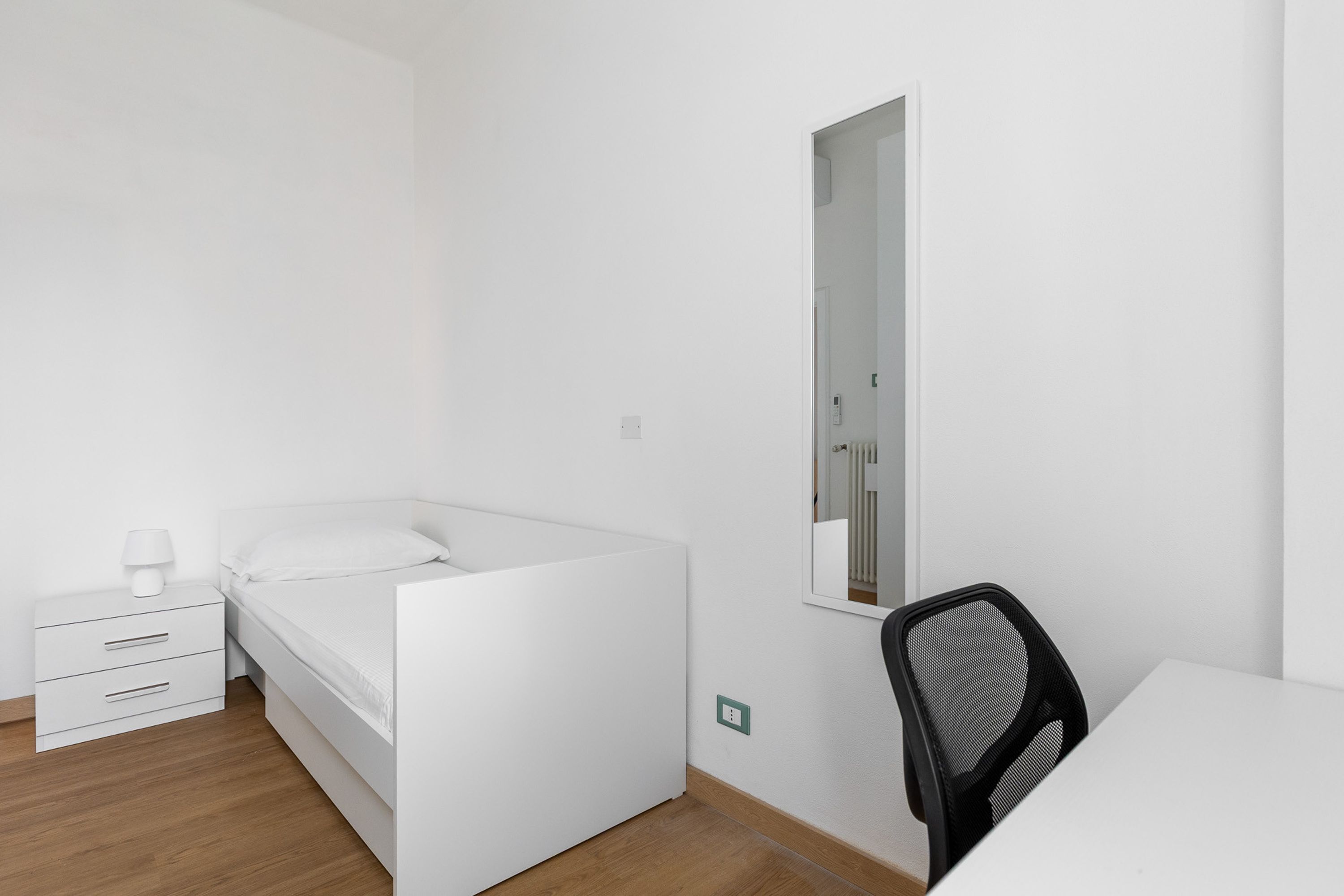A minimalist bedroom with a bed, a nightstand, a chair, and a mirror.