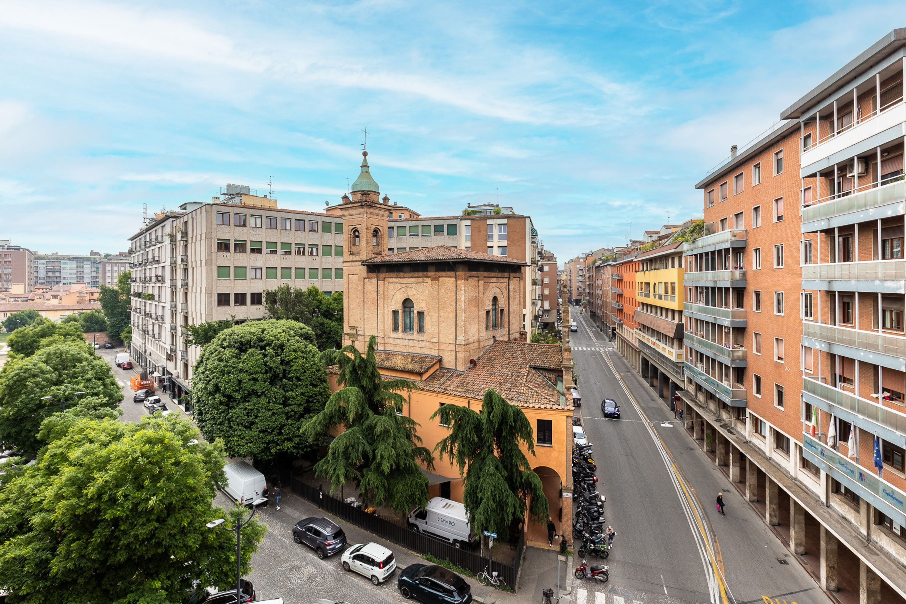 A cityscape with a mix of modern and historic buildings, a clear sky, and a street with parked cars and a few pedestrians.