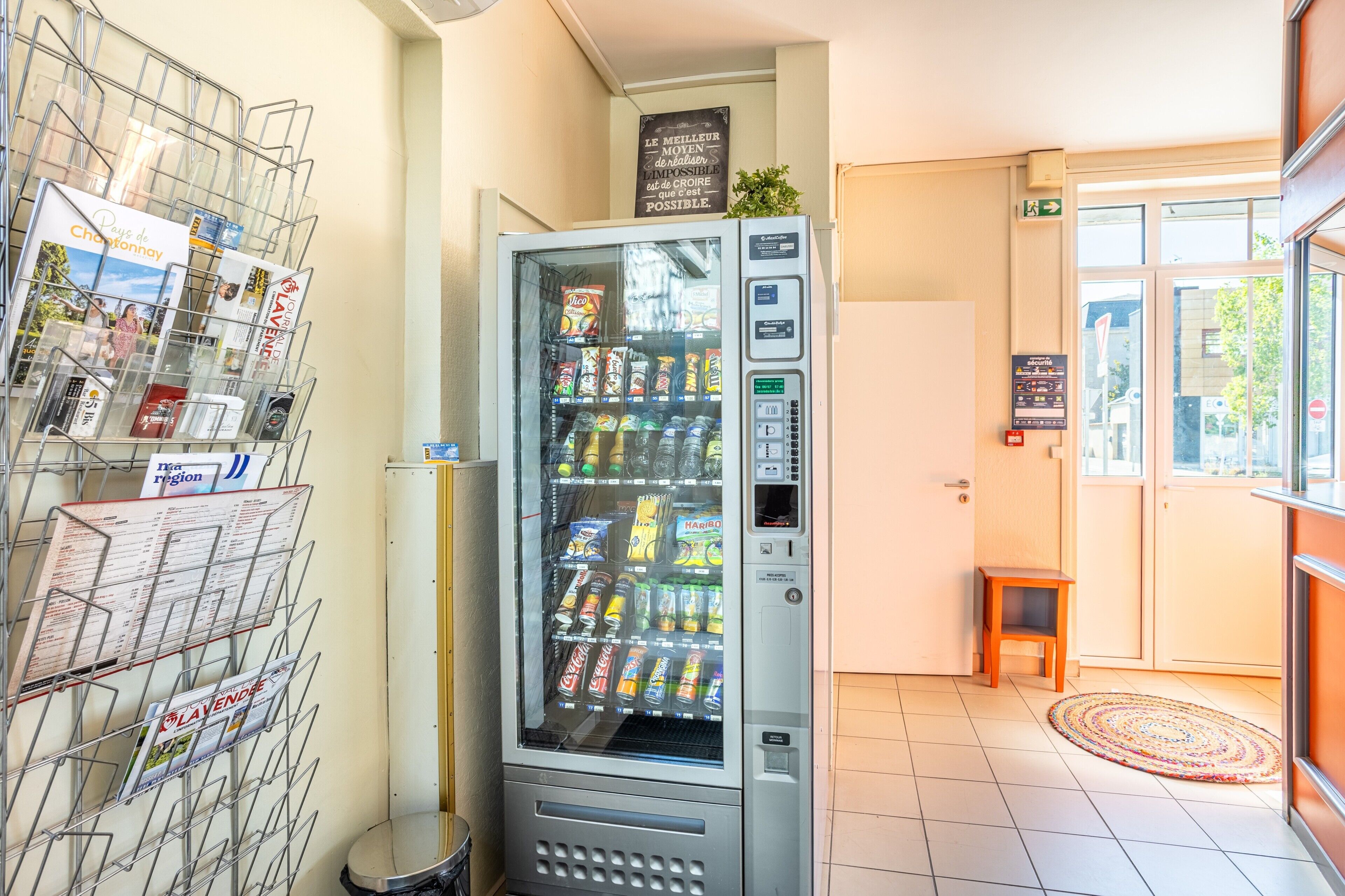 Un distributeur automatique de collations et de boissons, un présentoir à magazines et un petit tabouret en bois.